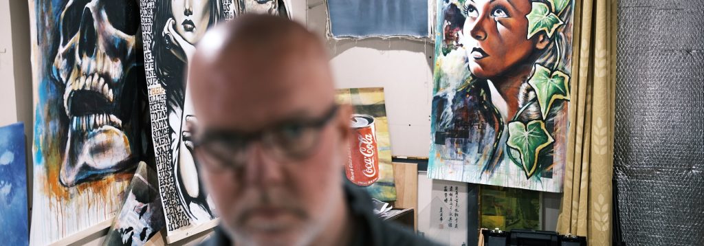 Blurred foreground portrait of Alex Loveless in the studio, with paintings and works in progress on the wall behind him.