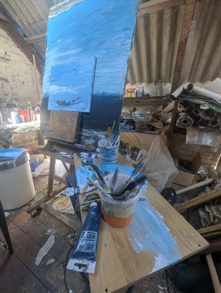 Studio view of blue oil paint, brushes, palette knives, and a landscape painting in progress.